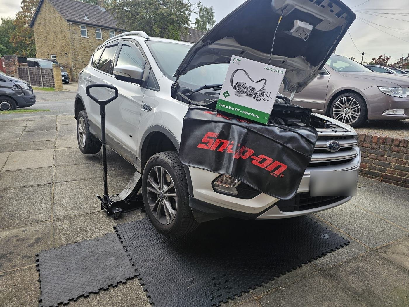 A timing belt kit and water pump box resting on a Ford with its bonnet open, on workshop mats