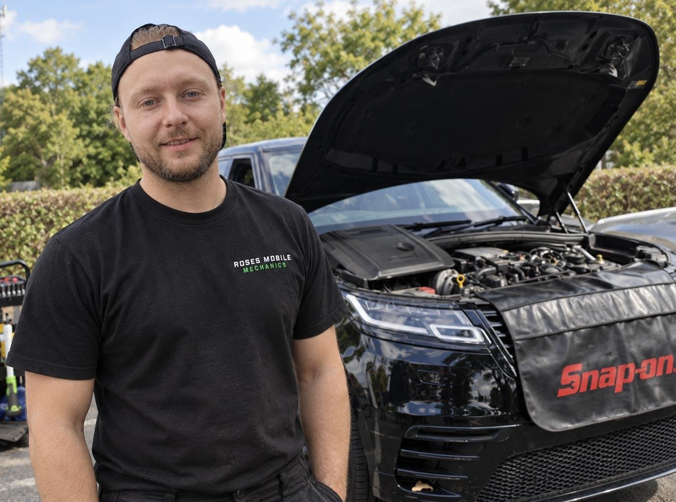 Seb Rose in a Roses Mobile Mechanics shirt, standing beside a car with its bonnet open